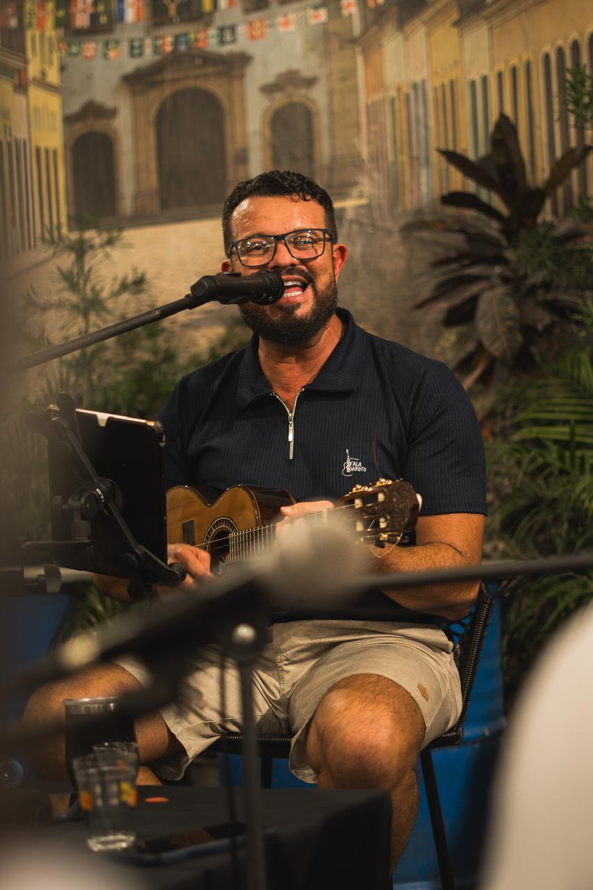 Vocalista do Fala Garoto, de óculos e camisa azul-marinho do grupo, cantando ao microfone com o cavaquinho no colo